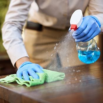 Person in blue gloves cleaning a wooden table with a green cloth and spray bottle