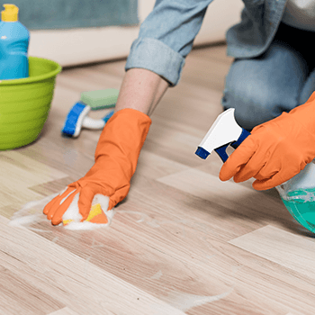 Person cleaning a wooden floor with a spray bottle and cloth, wearing orange gloves